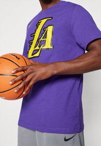 Fialové bavlněné tričko s žlutou grafikou "LA", nošené s šedými sportovními kraťasy. Míč na basketball je držen jednou rukou, přičemž je vidět texturovaný povrch.