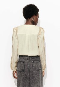 Blouse couleur crème avec un tissu texturé, des manches brodées et un dos plissé, associée à une jupe en denim foncé dotée de poches.