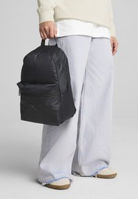 Mochila negra con diseño texturizado, bolsillo con cremallera en la parte frontal y logo de PUMA. El portador la sostiene por la asa superior, vistiendo unos jeans de corte holgado.
