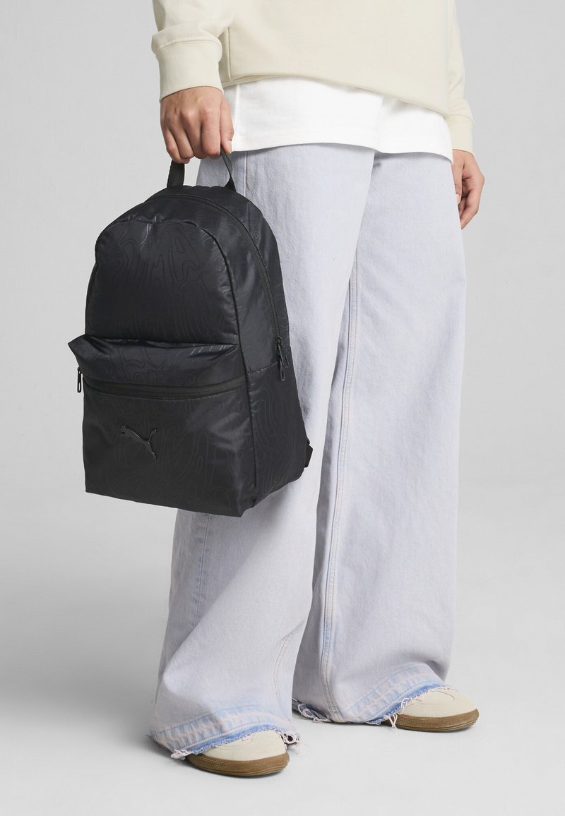 Mochila negra con diseño texturizado, bolsillo con cremallera en la parte frontal y logo de PUMA. El portador la sostiene por la asa superior, vistiendo unos jeans de corte holgado.