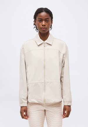 Rövid, fonott hajú nő világos bézs színű cipzáras dzsekit visel, amelynek rugalmas mandzsettái vannak, és mintás, világos nadrágot, egy egyszínű háttér előtt áll.