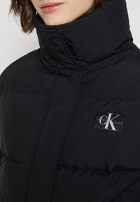 Sort pufferjakke med høj krave og kviltet tekstur. Har Calvin Klein Jeans-logo på venstre bryst. Minimalistisk design.