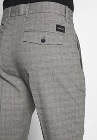 Pantalones de cuadros grises con dos bolsillos laterales y un bolsillo trasero con botón. La etiqueta muestra "Calvin Klein" en negro.