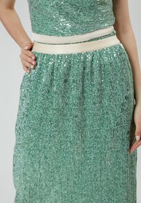 Vestido verde con lentejuelas de diseño texturizado y plisado. Presenta una amplia cinturilla en crema. La sección superior más corta crea un efecto de capas.