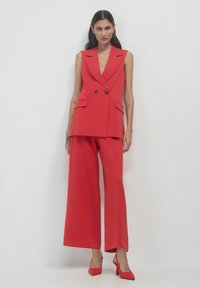 Gilet rosso su misura con doppi bottoni e tasche frontali, abbinato a pantaloni larghi rossi e scarpe con tacco abbinate. Texture liscia.