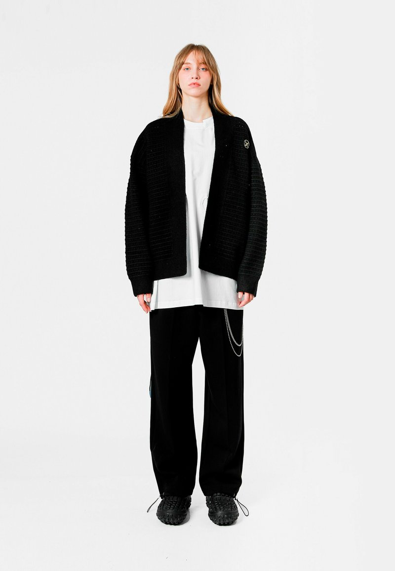 Cardigan negro texturizado de corte holgado, llevado sobre una camiseta blanca de gran tamaño. Pantalones amplios negros y calzado negro texturizado completan el look.
