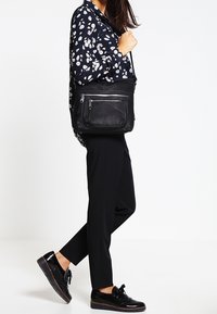 Donna con blusa nera a fiori, pantaloni neri e mocassini neri lucidi, che porta una borsa a tracolla in pelle nera con cerniere frontali.