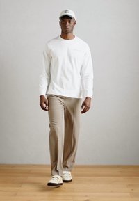 Bílá košile s dlouhým rukávem, béžové kalhoty a bílé boty s hnědými akcenty. Model v modré kšiltovce, stojící na jednobarevném pozadí.