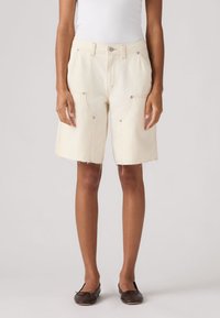 Γυναίκα που φοράει κρεμ τζιν σορτς με ψηλή μέση και ακατέργαστο τελείωμα, λευκό αμάνικο τοπ και σκούρα καφέ μπαλαρίνες, στέκεται σε ανοιχτό φόντο.