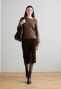 Pulover tricotat texturat de culoare maro, cu un croi lejer, asortat cu o fustă tip creion de culoare maron închis, cu o slit lateral. Modelul ține o geantă tote neagră.