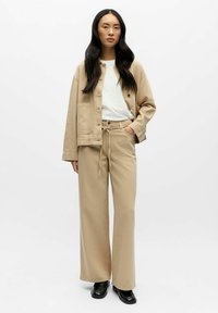 Bēša pārmēra izmēra jaka ar pogām, balta T-krekls un bēšas augsta vidukļa bikses. Melni apavi noslēdz izskatu. Gluda auduma, brīvāka piegriezuma.