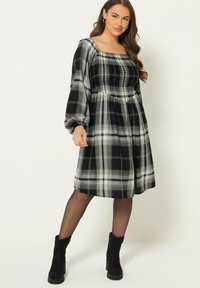 Mustat ja valkoiset ruudulliset mekko, jossa on neliön muotoinen pääntie ja puffihuput. Kankaat ovat kevyet ja sileät. Mustat nilkkurit täydentävät asun.