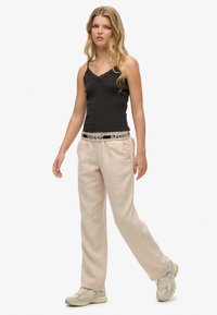Camisola nera con pizzo, pantaloni beige a gamba larga con texture, e scarpe da ginnastica color neutro. Modella in una posa rilassata.