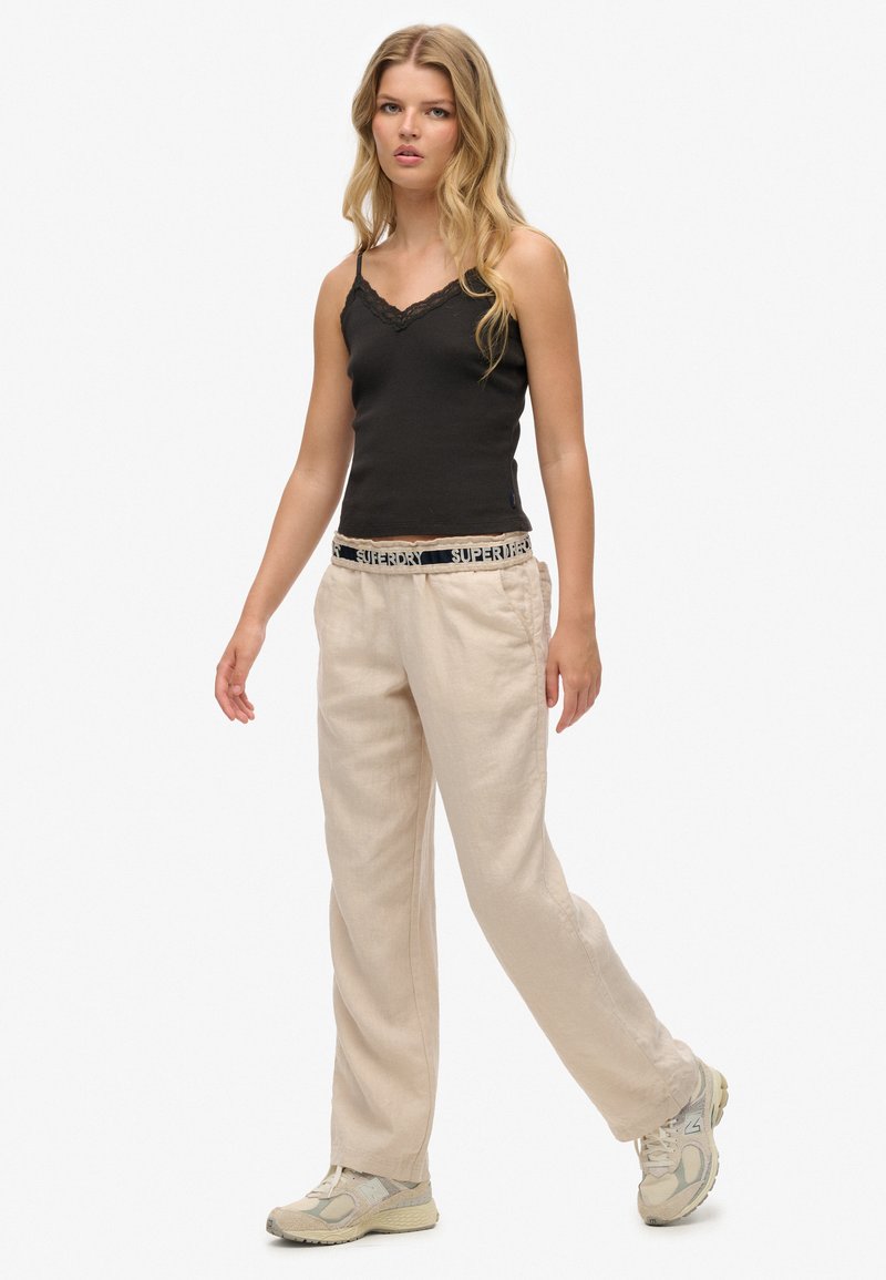 Camisola nera con pizzo, pantaloni beige a gamba larga con texture, e scarpe da ginnastica color neutro. Modella in una posa rilassata.
