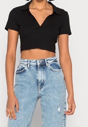 Femme portant un polo court noir à manches courtes et un jean taille haute délavé bleu clair, debout les mains détendues.