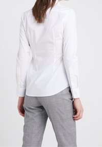 White long-sleeve shirt with a fitted silhouette, back seam details, and buttoned cuffs, paired with gray checked tailored pants.