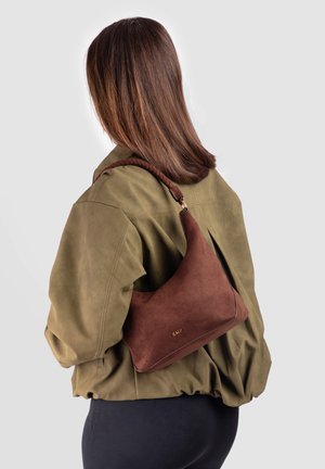 Γυναίκα με καστανά μαλλιά μέχρι τους ώμους, που φοράει ελιά μπουφάν και κρατάει μια καφέ σουέντ τσάντα στον ώμο της μπροστά σε απλό φόντο.