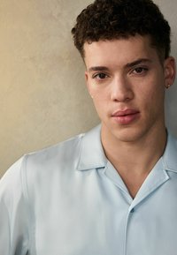 Camisa de manga corta azul claro con una textura suave y cuello abotonado. El sujeto tiene el cabello corto y rizado, y está posicionado frente a un fondo neutro.