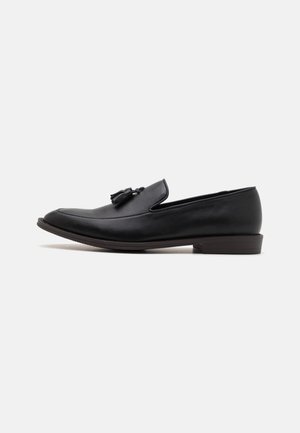 Mocassin à enfiler en cuir noir avec détail à glands et petit talon empilé, vu de profil sur fond blanc.