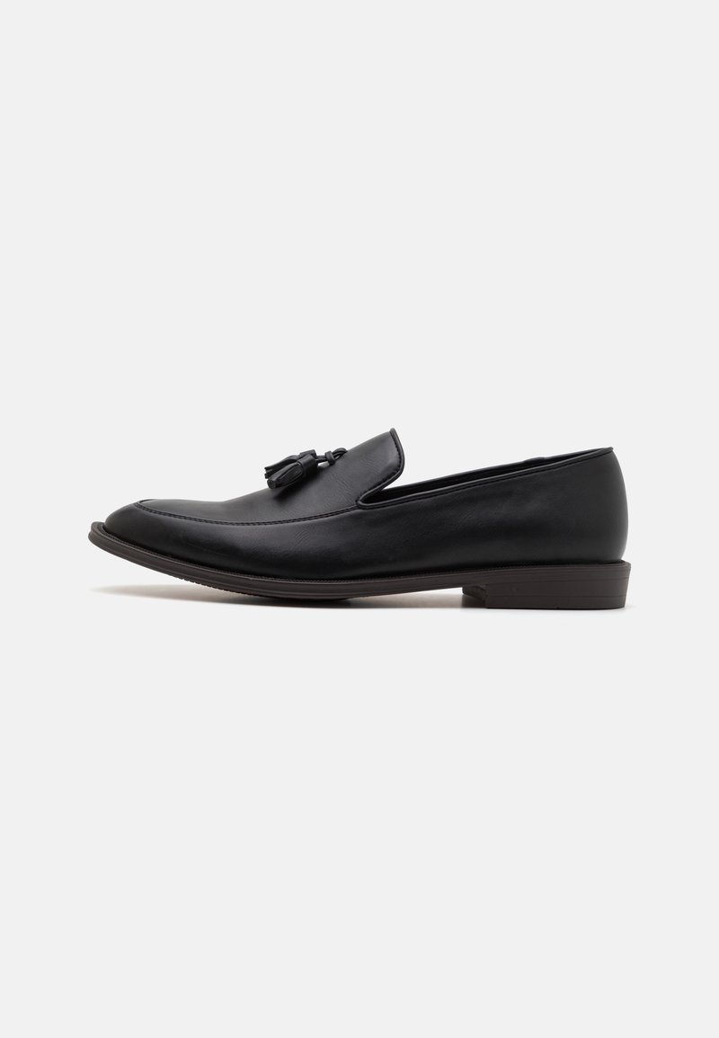 Mocassin à enfiler en cuir noir avec détail à glands et petit talon empilé, vu de profil sur fond blanc.