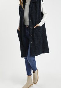 Granatowa, teksturowana kamizelka z kapturem, przednimi kieszeniami i zapięciem na napy. Noszona na jasnym, długim rękawie i niebieskich dżinsach.