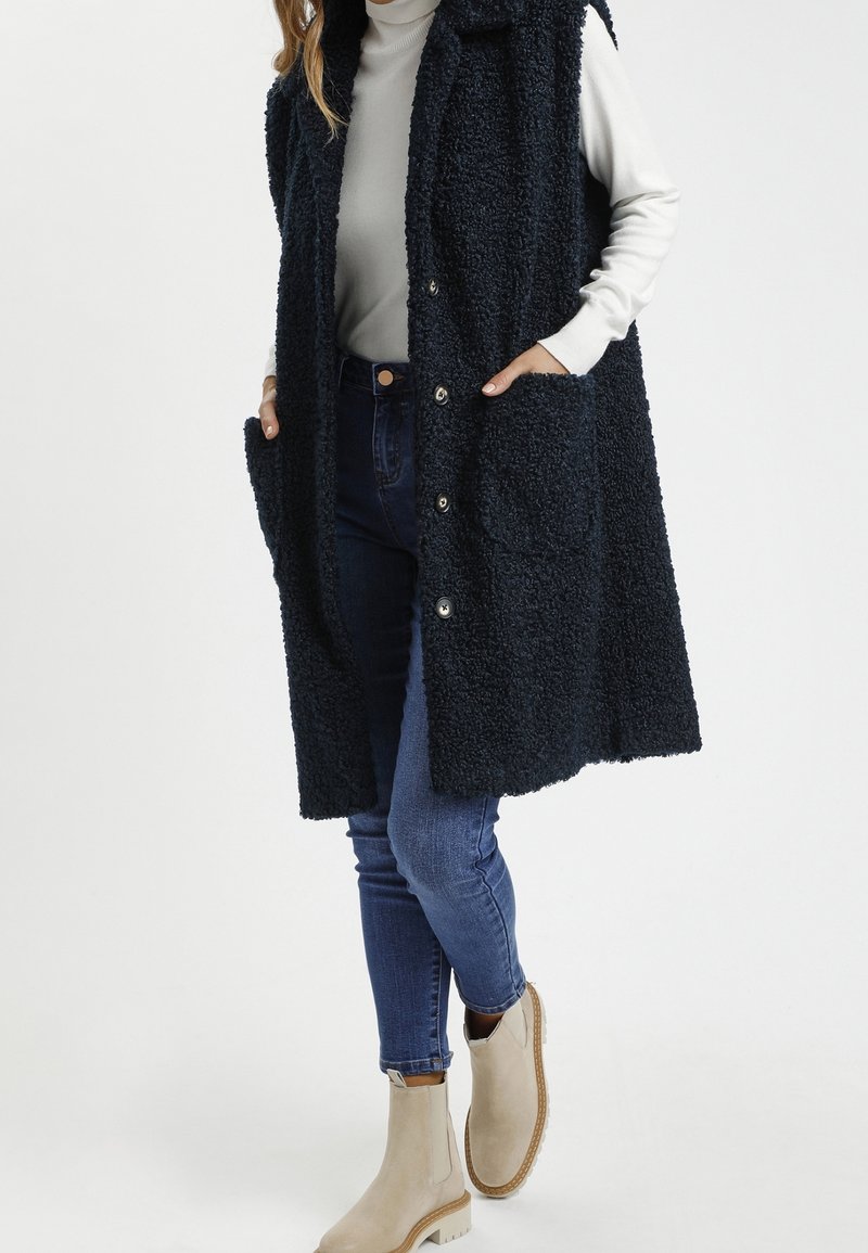 Granatowa, teksturowana kamizelka z kapturem, przednimi kieszeniami i zapięciem na napy. Noszona na jasnym, długim rękawie i niebieskich dżinsach.