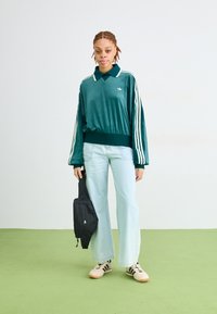 Osoba u tirkiznoj Adidas dukserici, svijetloplavim širokim trapericama, bež tenisicama, drži crnu torbu pred jednostavnom bijelom i zelenom pozadinom.