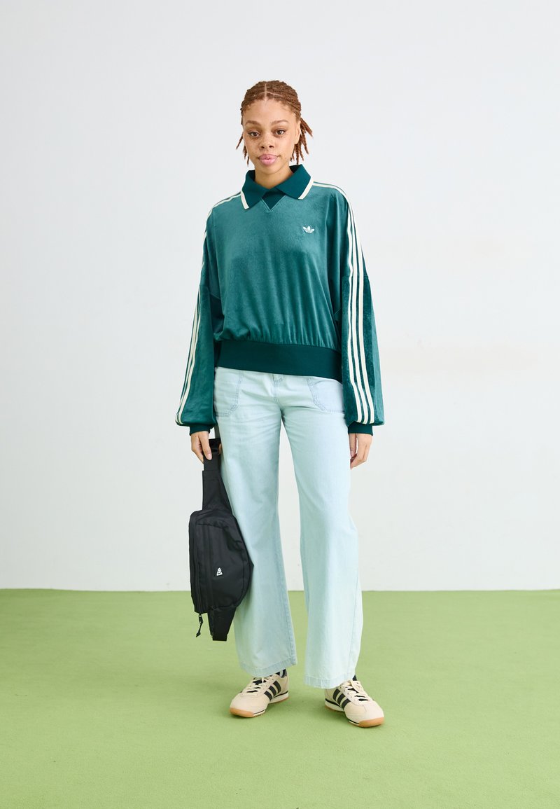 Persoon in een teal Adidas sweatshirt, lichtblauwe wijde jeans, beige sneakers, met een zwarte tas tegen een effen witte en groene achtergrond.
