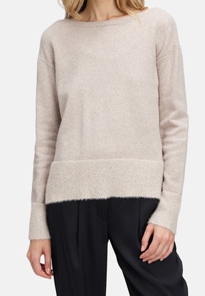 Vrouw draagt een lichtbeige gebreide trui met geribbelde manchetten en zoom, gecombineerd met loszittende zwarte broek.