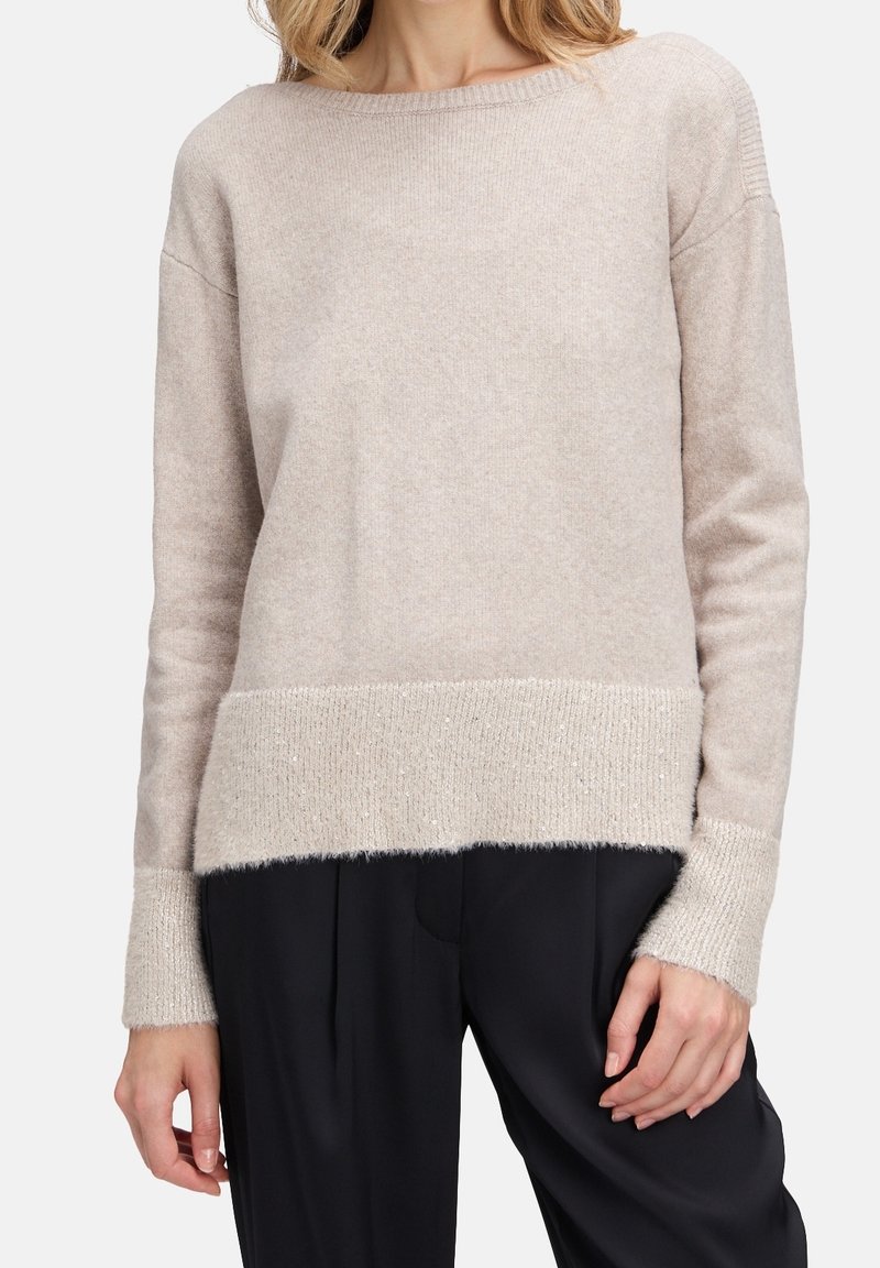 Femme portant un pull en maille beige clair avec poignets et ourlet côtelés, associé à un pantalon noir ample.