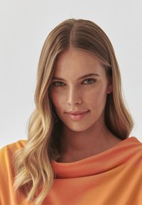 Detailní záběr na osobu s dlouhými, vlnitými blond vlasy, která má na sobě hladký, oranžový top s širokým výstřihem na světlem pozadí.