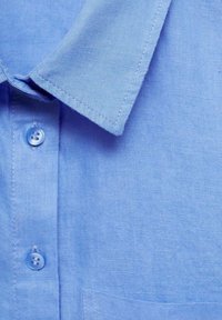 Nahaufnahme des hellblauen Hemdkragens, zwei Knöpfe und strukturierter Stoff mit sichtbaren Nähdetails.