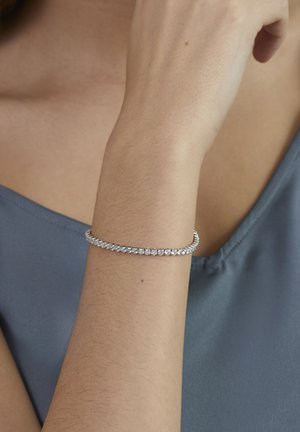 Frau trägt ein silbernes Tennisarmband mit kleinen Diamanten am Handgelenk, gekleidet in einem blauen ärmellosen Top.