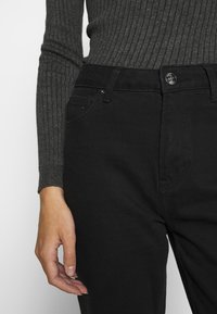 Jeans de mezclilla negros con cintura alta, con bolsillos frontales y cierre de botón. Parte de una camiseta de manga larga de color gris oscuro y acanalada es visible.