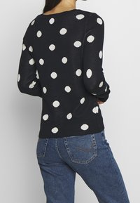 Mustapaita, jossa on valkoisia pilkkuja, ribatut kaula-aukko ja hihansuut, vartalonmyötäinen muoto ja pehmeä rakenne, yhdistettynä sinisiin farkkuihin.