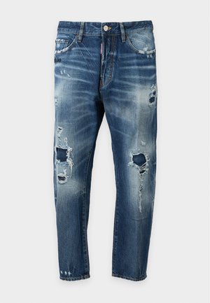 Jeune homme portant une chemise blanche à manches courtes, jean bleu déchiré, bottes marron, colliers croix, portant un sac à dos blanc, debout avec la main dans la poche.