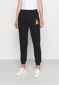 Svarta sweatpants med ribbad midja, uppkavlade anklar och en guld broderad design på låret. Bärs med vita sneakers.