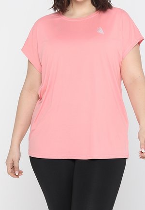 Vrouw draagt een loszittende roze top met korte mouwen en een klein wit logo op de borst, gecombineerd met strakke zwarte broek.