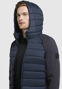 Gilet matelassé à capuche bleu marine avec texture quiltée, fermeture éclair et accents en tissu noir. Présente un patch rond avec un logo sur l'épaule.