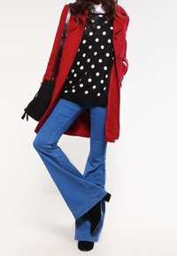 Abrigo largo rojo, suéter negro con lunares, y jeans acampanados azules. La textura varía; el suéter es de punto y los jeans son de mezclilla. Incluye un bolso negro.
