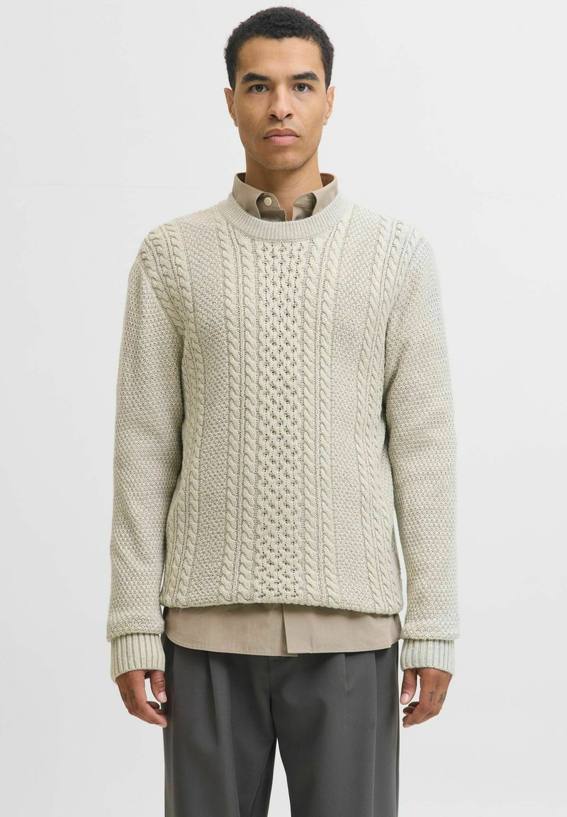 Pull en tricot beige clair avec des motifs en torsades et texturés, à manches longues et un col rond côtelé. Porté par-dessus une chemise à col.
