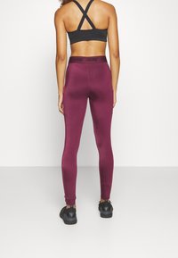 Bordowe legginsy o gładkiej teksturze i wysokim pasie z czarnym logo, połączone z czarnymi butami z logiem na pięcie.