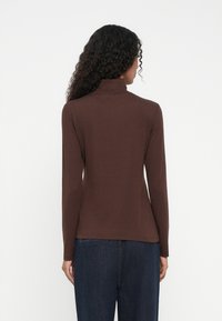 Vrouw met lang krullend haar, gekleed in een aansluitende bruine coltrui en donkerblauwe jeans, staand met haar rug naar de camera tegen een effen achtergrond.