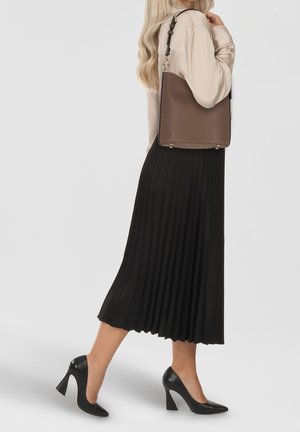 Femme portant une jupe midi plissée noire, des talons hauts noirs, un haut beige à manches longues, avec un sac à main en cuir marron porté à l'épaule.