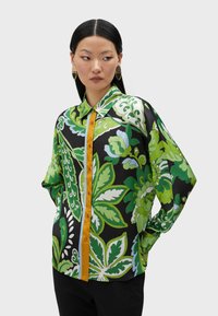 Mångfärgad blus med stora gröna blommönster på en svart bakgrund, med en gul knappslå och en avslappnad passform.