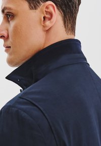 Granatowa kurtka z wysokim kołnierzem i żebrowanymi akcentami, wykonana z gładkiego materiału i dopasowanego kroju, widoczna z boku.