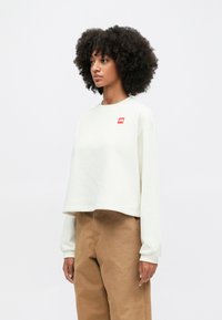 Κοντό, κρεμ φούτερ με μακριά μανίκια και κόκκινο λογότυπο στο στήθος. Λείο ύφασμα, χαλαρή εφαρμογή, συνδυασμένο με καφέ παντελόνι.