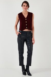 Kvinde iført ærmeløs bordeaux vest med guldknapper, sorte læderbukser og sorte ankelstøvler, stående med den ene hånd i lommen.