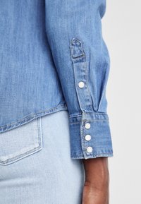 Poignet de manche d'une chemise en denim bleu avec quatre boutons blancs, portée par une personne à la peau foncée, associée à un jean en denim bleu clair avec poche visible.