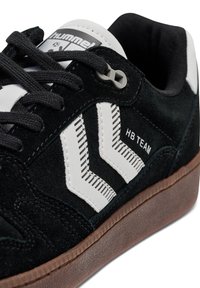 Schwarze Wildleder-Sneaker mit weißen Lederakzenten, diagonal gestreiftem Design, schwarzen Schnürsenkeln und brauner Gummisohle. Logo auf der Zunge sichtbar.